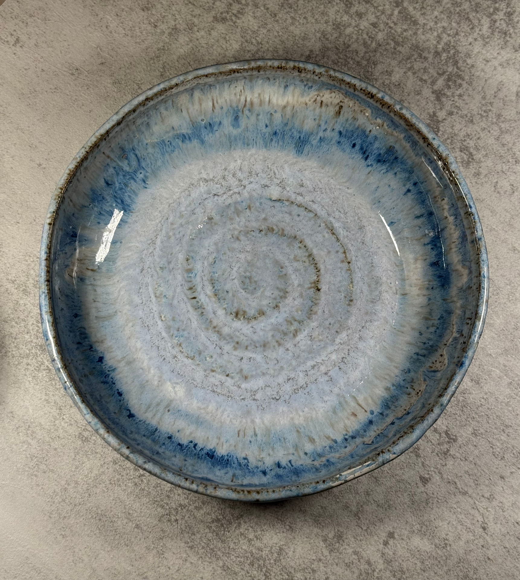 Overhead view of a shallow handmade bowl with a pale center fading through cerulean into deep cobalt blue, a toasted brown rim, and a spiral swirl at the base