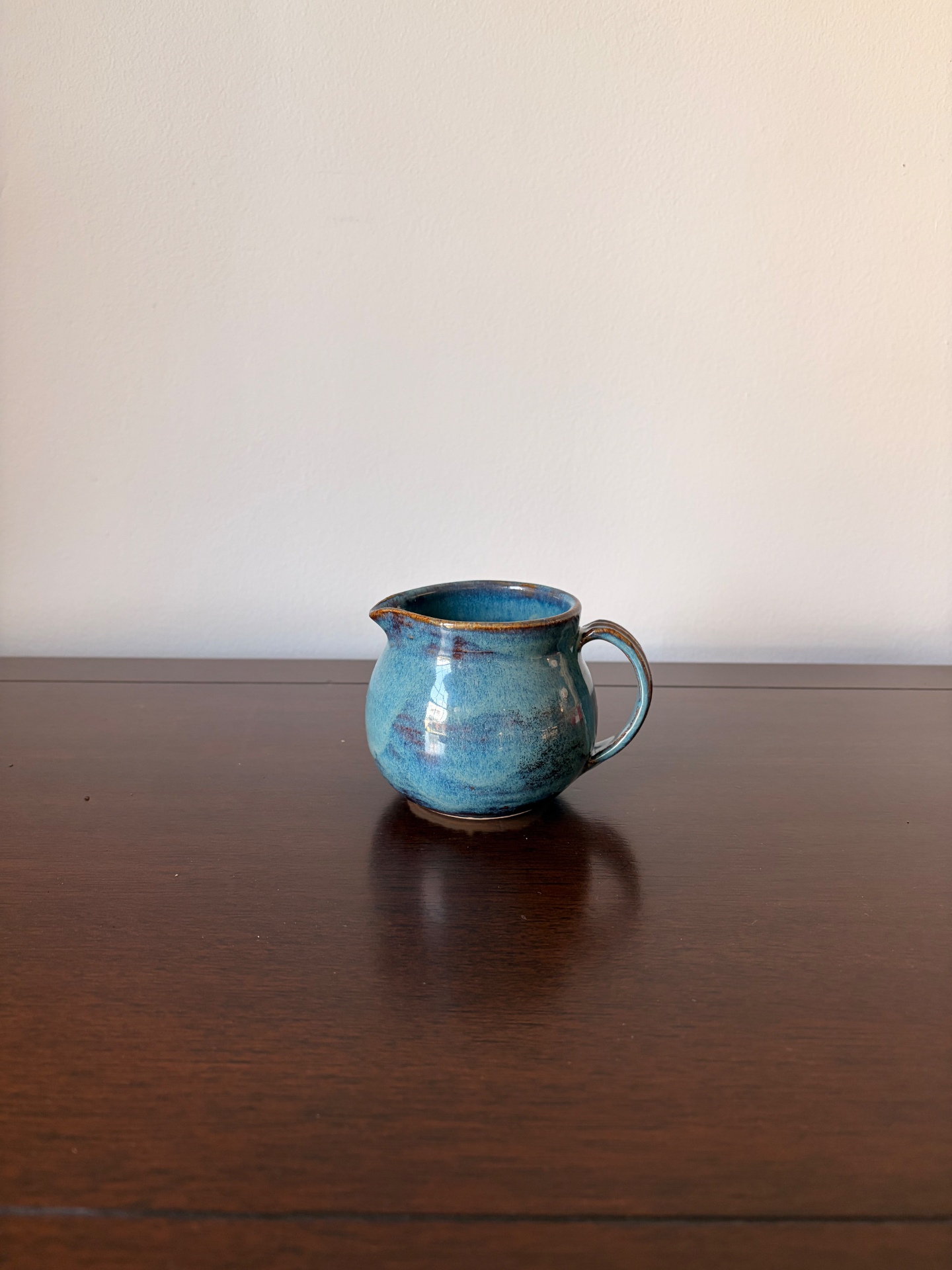 Round ceramic pitcher with handle in mottled ocean blue glaze with rust accents on a dark wood table